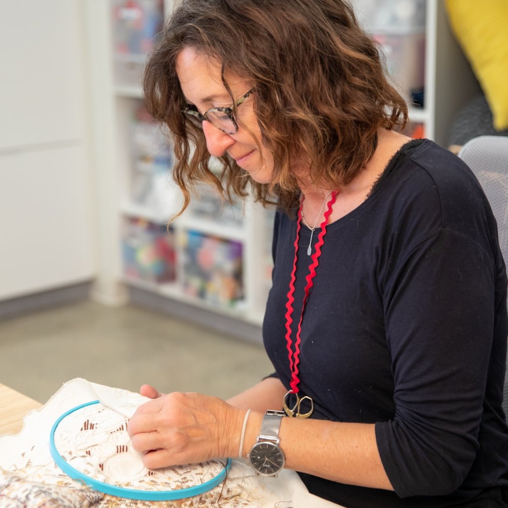 A woman sitting at a table with an embroidery hoop in front of her.