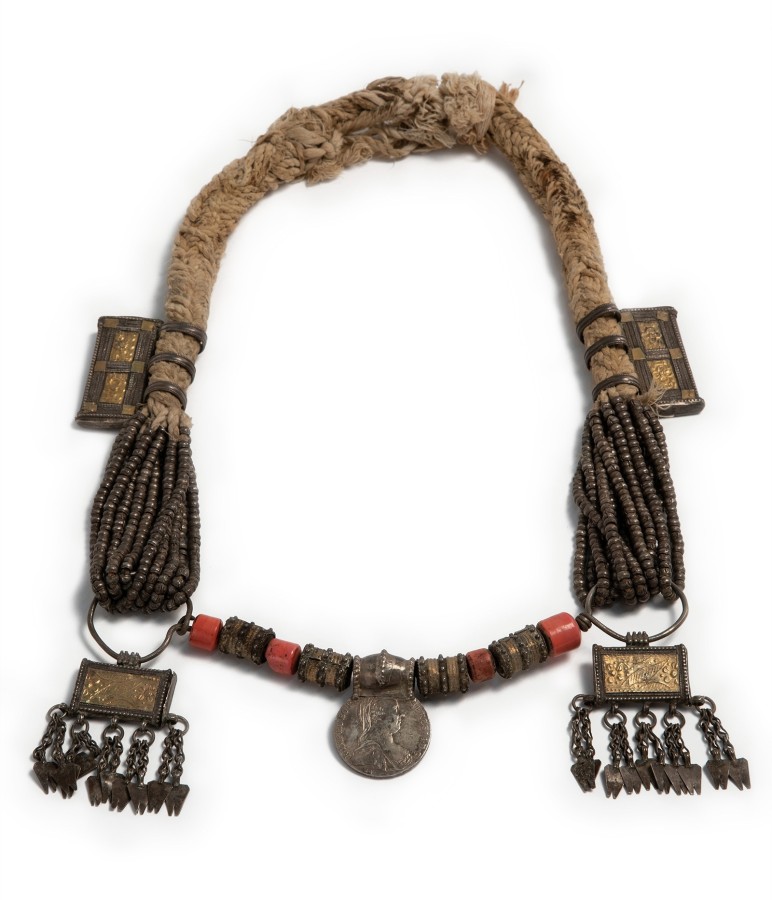 This necklace is a mix of materials such as cotton rope, glass and silver. In the center is a silver medallion and to either side of the medallion are glass beads. Some of the glass beads are made to look like coral..