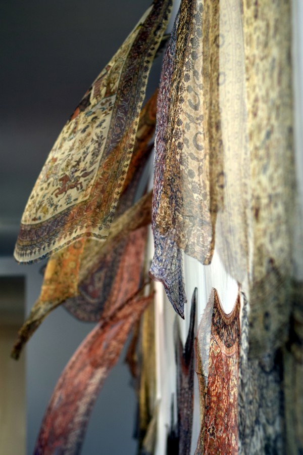 Shirin Towfiq, "Thinking About Migration", 20 digital prints on gauze with fan, 15’ x 10’ installation. Courtesy of the Artist. 

Image: side view of very lightweight textiles installed on a wall floating, appearing to be floating. On the textiles are intricate geometric patterns in creams, blues, reds, and oranges.