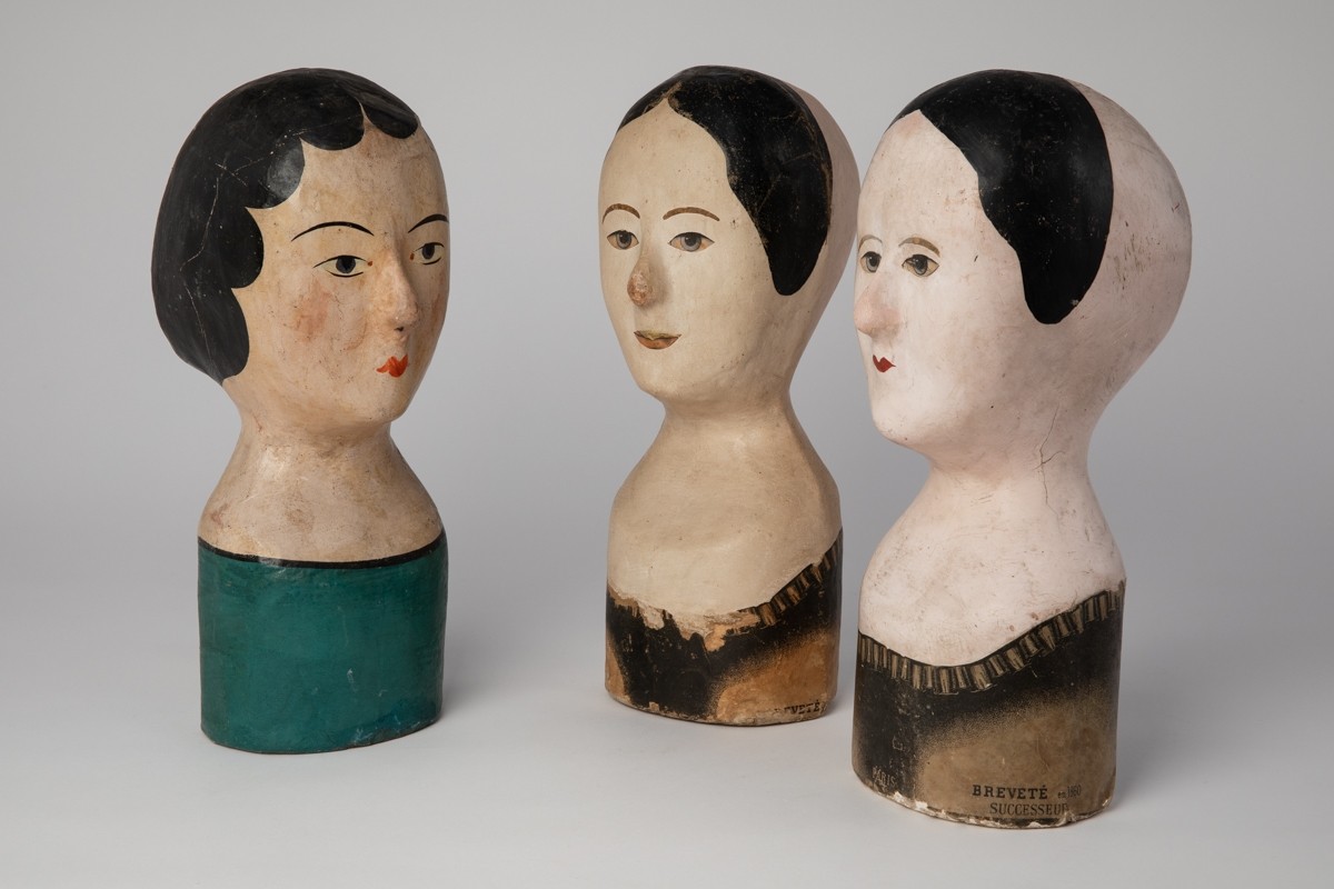 There are three milliner's store head forms. The two on the right face the one on the left.  The two on the right have short black hair. The one on the left has long black hair. They are all light complexioned.