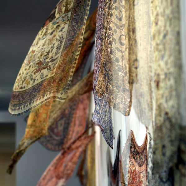 Thinking About Migration", 20 digital prints on gauze with fan, 15’ x 10’ installation. Courtesy of the Artist. 
Image: side view of very lightweight textiles installed on a wall floating, appearing to be floating. On the textiles are intricate geometric patterns in creams, blues, reds, and oranges.